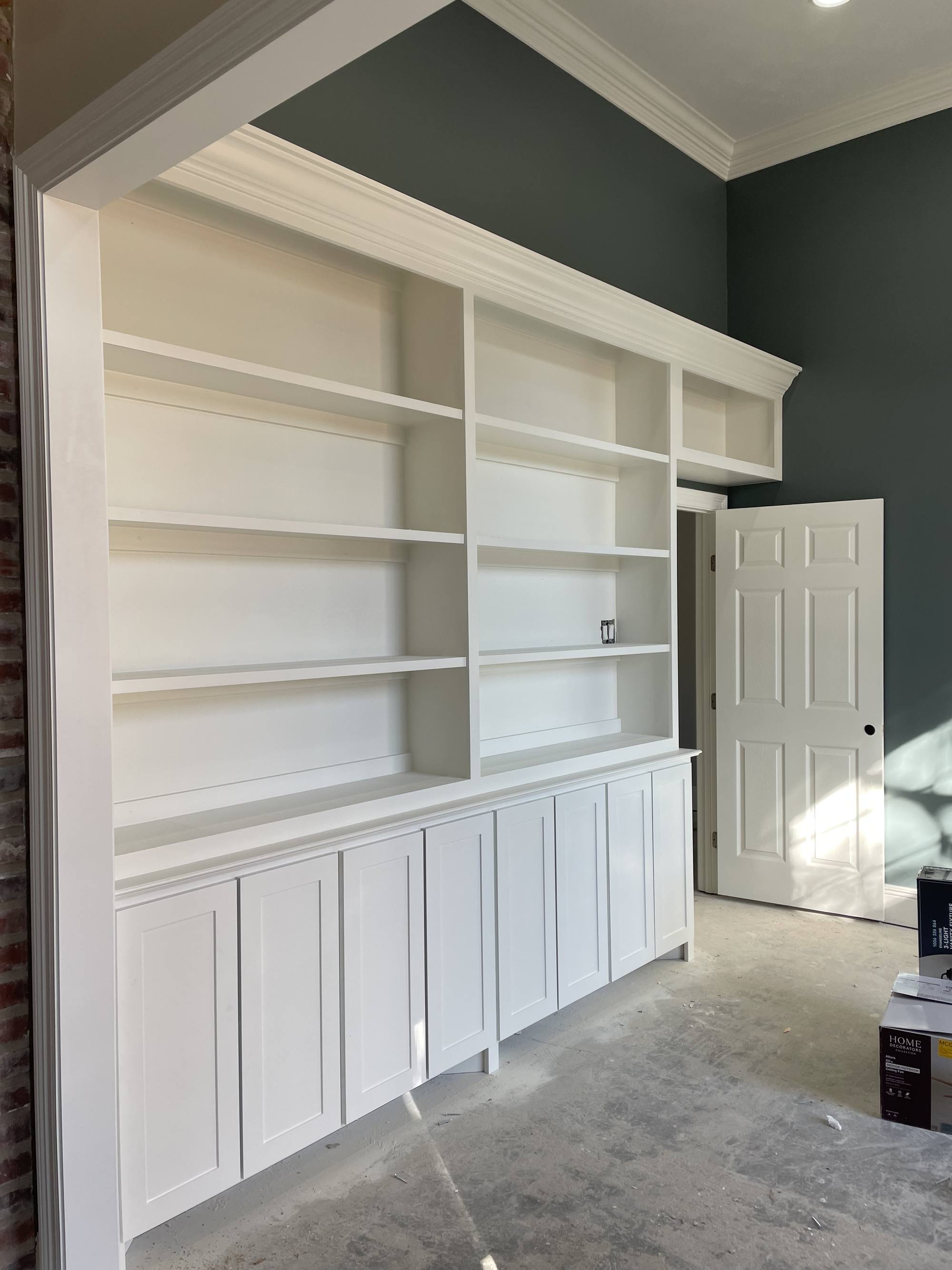 Floor-to-ceiling white built-in bookcase with shaker base cabinets against a dark green wall